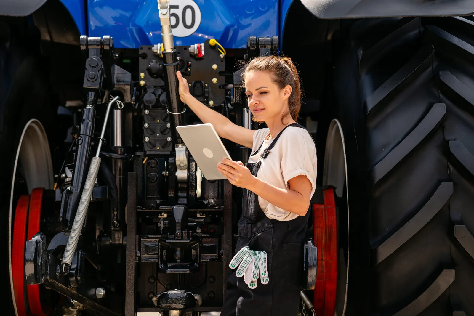 Person kontrollerer koblinger bak på en blå traktor med nettbrett